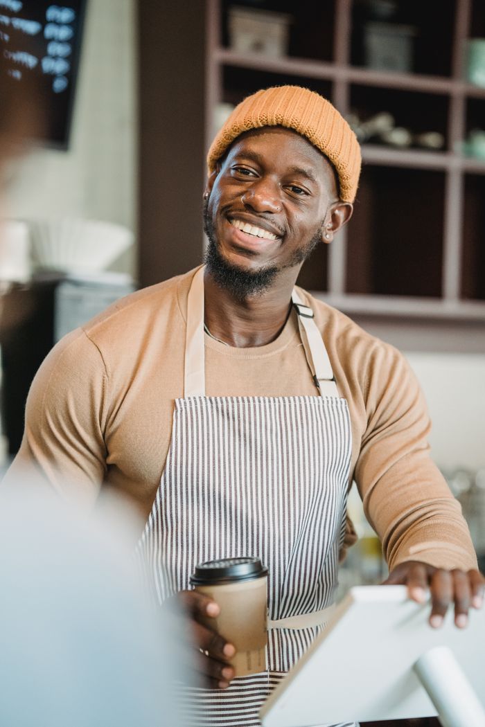 A cheerful barista in a beanie and apron serves coffee at a cozy cafe.