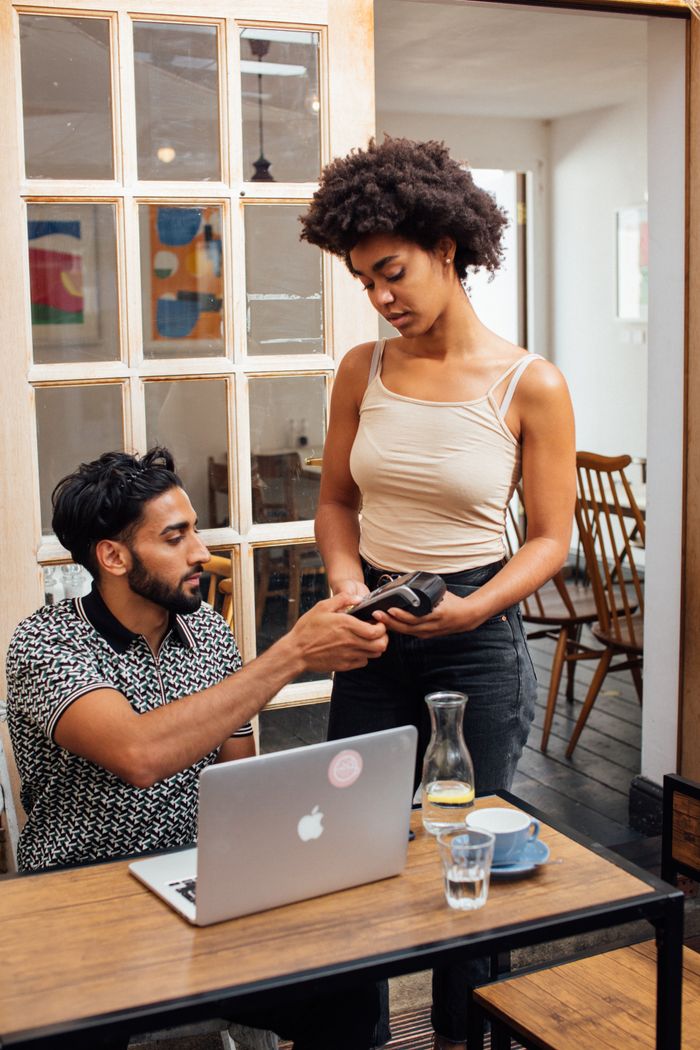 Man at laptop discusses payment with woman using a card reader in a cozy cafe.